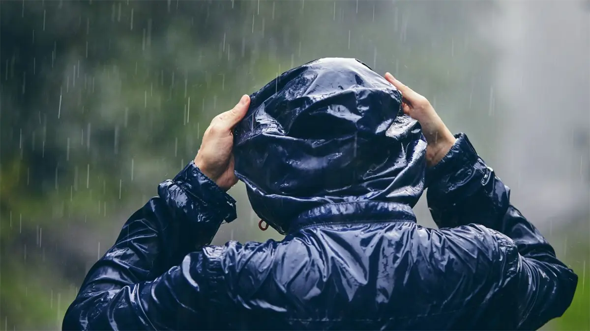 La imagen muestra a una persona de espaldas, vestida con una chaqueta impermeable oscura con capucha, bajo una lluvia intensa.