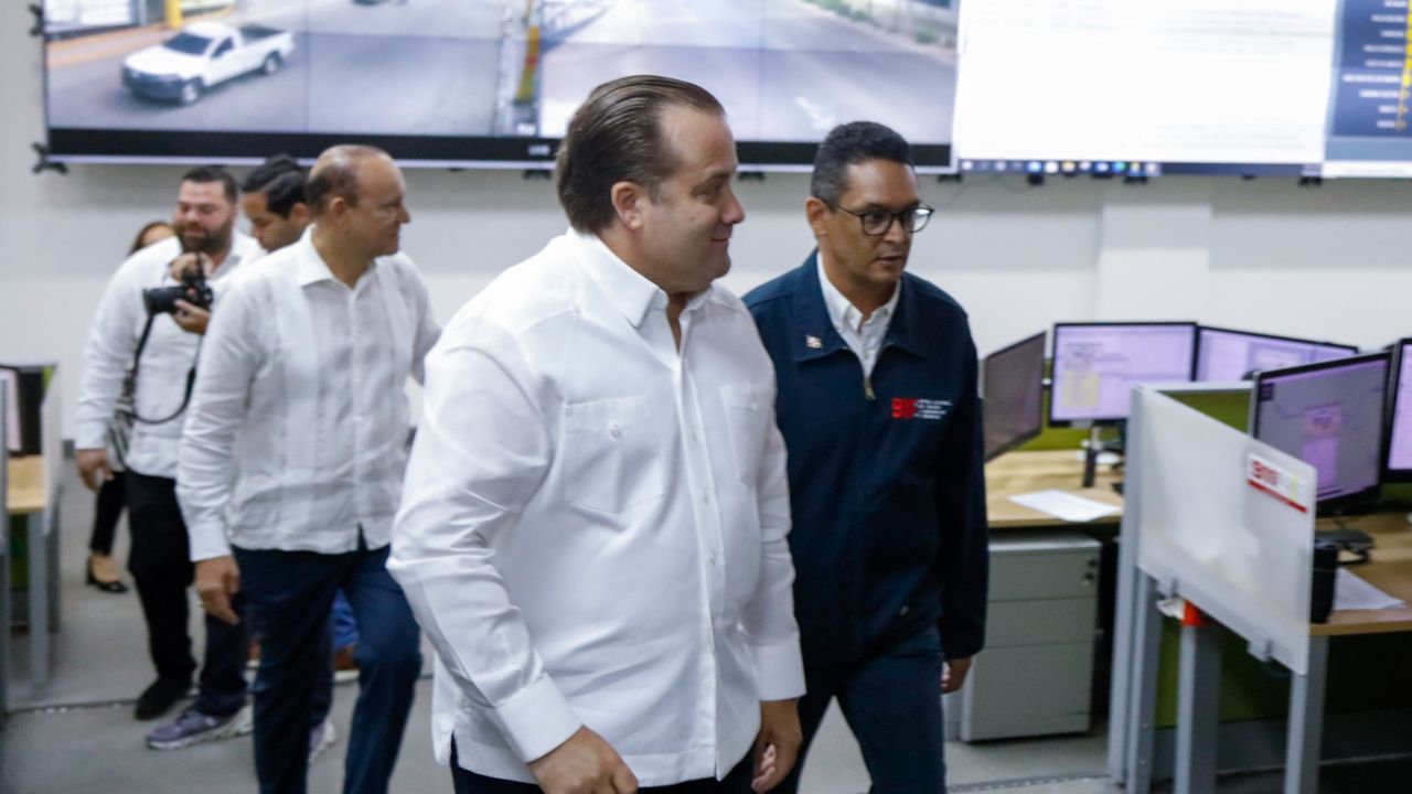 Personas caminan en sala con monitores y cámaras.