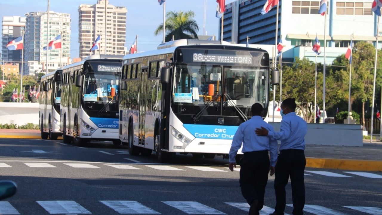 Autobuses circulando por avenida con banderas y edificios.