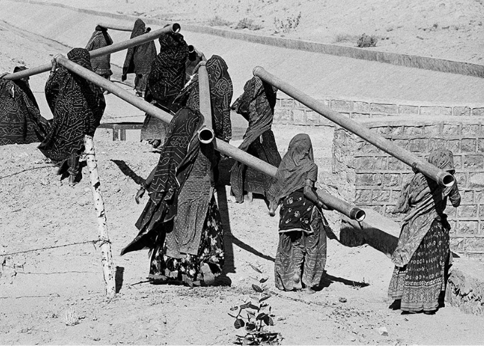 Mujeres llevando tubos por camino arenoso.