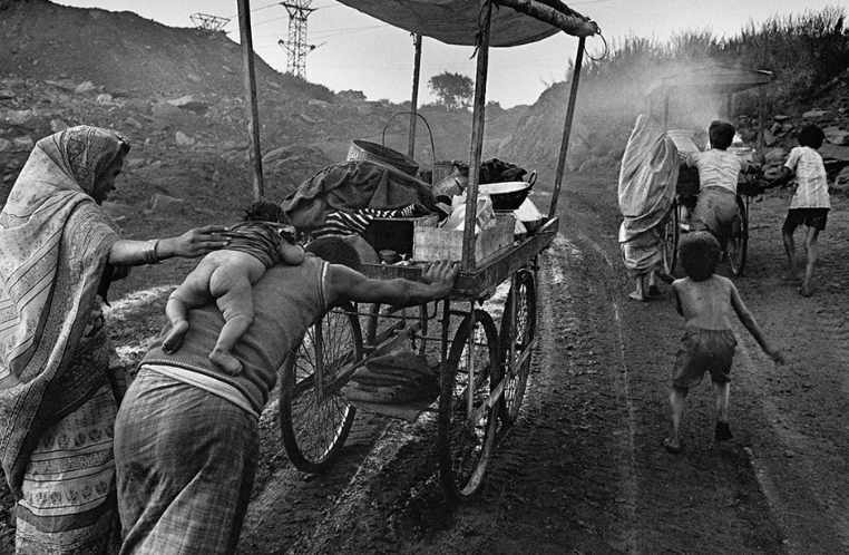 Familia empuja carrito por camino rural.