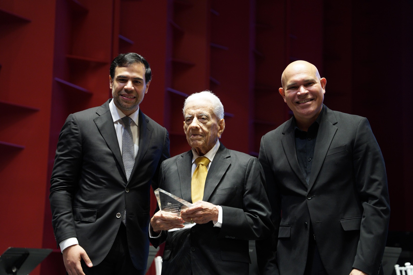 Tres hombres en trajes formales, uno sostiene premio.
